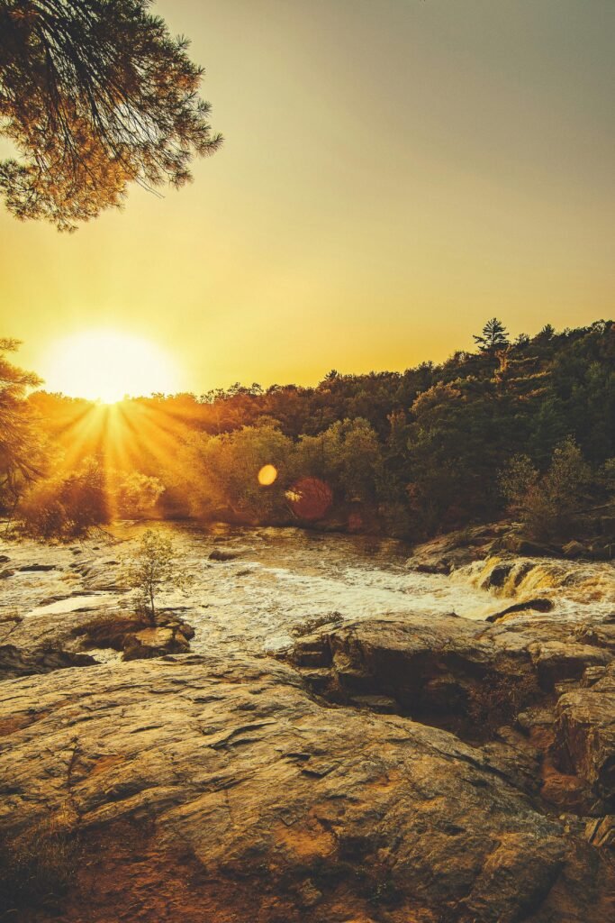pexels-photo-1420440-1420440 Stunning sunset over a rocky river surrounded by lush greenery and sunbeams creating a serene landscape.